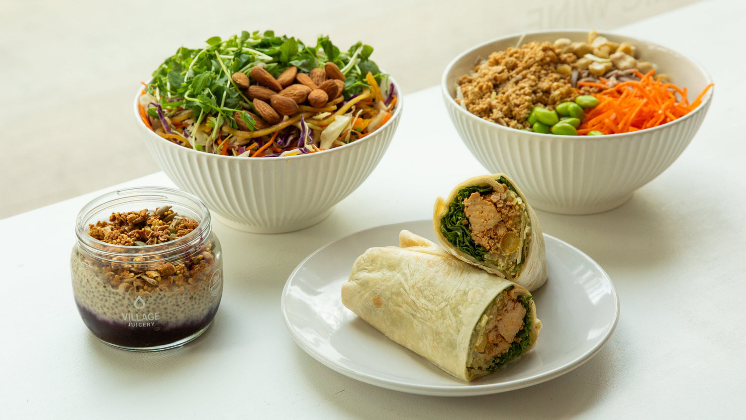 Assorted food items including a wrap, bowls of salad and rice, and a jar on a white surface.